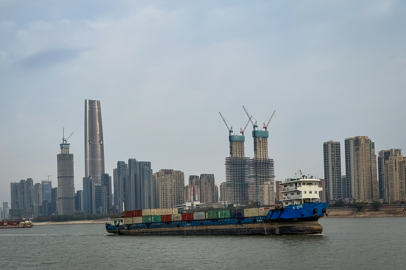 A cargo ship carrying containers sails along the Yangtze River, with Wuhan’s rapidly developing skyline and high-rise construction projects in the background. The city and its factories play a vital role in global trade and logistics