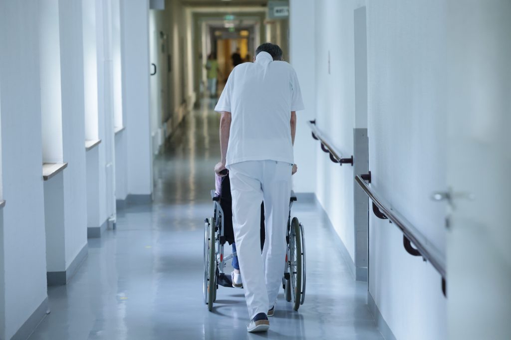 Image of a hospital hallway with a patient being wheeled away in a wheelchair.